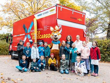 Grundschulklasse steht mit Maskottchen Blicki, Frau Humbaur und Frau Friedrichs vor einem LKW auf dem Schulhof | © Humbaur GmbH