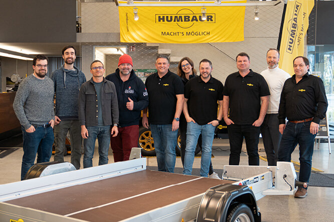 Humbaur Mitarbeitende und der Spanngurt COACH stehen nebeneinander vor einem Anhänger | © Humbaur GmbH